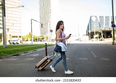 Stylish Caucasian Woman Using Mobile Phone While Pulling Rolling Suitcase On City Street