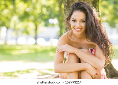 Stylish cheerful brunette sitting under a tree in a park on a sunny day