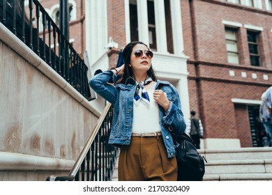 Stylish Chinese Lady Hand Combing Hair Is Standing On Outdoor Staircase With Metal Handrail. Modern Millennial Woman Is Looking Into Distance From A High Place, Background Red Brick Building