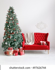 Stylish Christmas Interior With An Elegant Red Sofa. Comfort Home. Armchair With Fabric Upholstery. Christmas Tree With Presents Underneath In Living Room