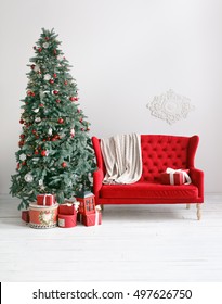 Stylish Christmas Interior With An Elegant Red Sofa. Comfort Home. Armchair With Fabric Upholstery. Christmas Tree With Presents Underneath In Living Room