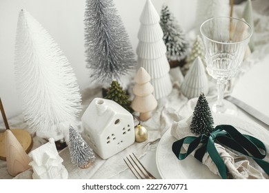 Stylish Christmas table setting. Festive napkin with ribbon and bell on plate, vintage cutlery, wineglass, modern christmas trees and houses on white rustic table. Holiday brunch - Powered by Shutterstock