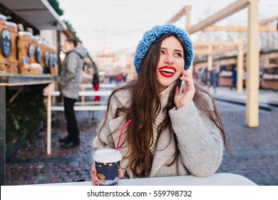 Stylish City Joyful Young Woman Sitting In Europe Modern City Centre, Wearing Winter Woolen Sweater, Blue Hat. Speaking On Phone, Smiling, Red Lipstick. Place For Text