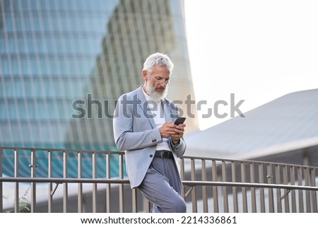 Stylish classy older mature adult professional business man, senior old bearded businessman executive holding smartphone, using mobile tech on cell phone standing outside big city office buildings.