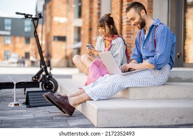 Stylish colleagues work on phone and laptop computer, charging them from portable solar panel, while sitting with electric scooter on a street at office district. Concept of sustainable lifestyle - Powered by Shutterstock