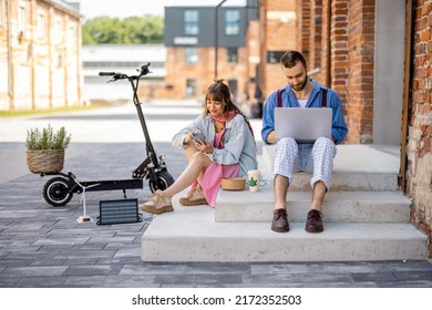 Stylish colleagues work on phone and laptop computer, charging them from portable solar panel, while sitting with electric scooter on a street at office district. Concept of sustainable lifestyle - Powered by Shutterstock