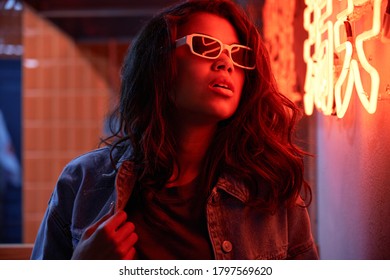 Stylish Cool Fashion Funky African American Young Beautiful Hipster Girl Mixed Race Model Wears Trendy Glasses Posing Near Red Neon Sign In Night Club. 80s 90s Gen Z Retro Style Party Nightclub Event.