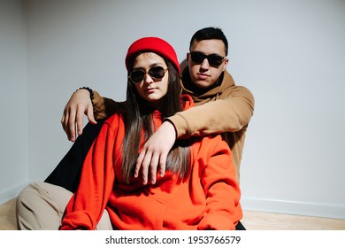 Stylish cool looking young couple in sunglasses and hoodies sitting on the floor on behind the other. In front of the white wall. Posing, trying to look cool.