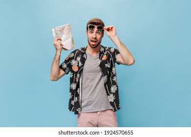 Stylish cool young man with beard in sunglasses and summer clothes looks at camera in shock and holds map on blue background..