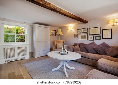 Stylish cottage living room containing sofa with large cushions, chic simple decor, numerous picture frames, coffee table, cupboard window and exposed beam