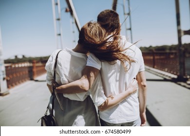 stylish couple in love hugging, back view with windy hair, on bridge in the summer city. modern woman and man in fashionable white clothes embracing at the river, romantic moment