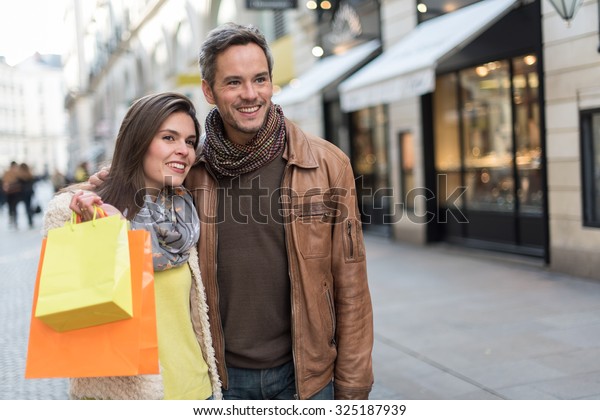 Stylish\
couple standing in a cobbled car-free street. The grey hair man\
with beard is wearing a brown leather coat and the woman a yellow\
top and two shopping bags, they also have\
scarfs