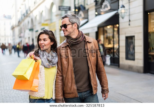 Stylish couple walking in a cobbled car-free\
street. The grey hair man with beard is wearing sunglasses and a\
brown leather coat and the woman a yellow top and two shopping\
bags, they also have\
scarfs