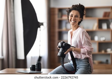 Stylish creative brunette attractive young woman in casual professional photographer or blogger sitting at table at home studio or home office holding camera and smiling indoors, copy space