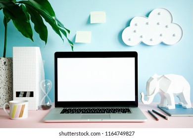 Stylish and creative desk with laptop mock up screen, office accessories, tropical leaf, cup of coffee, elephant figures and hanging cloud. Blue background wall. Design home office interior. 
