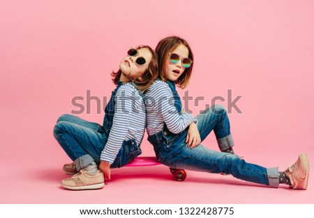 Stylish cute girls in fashion clothes on a skateboard on pink background. Twin girl child with skateboard.