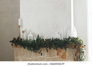 Stylish decorated christmas fireplace. Modern christmas trees, houses, pine cones and spruce branches on fireplace mantel with brass bells and ribbon. Christmas scandinavian living room decor - Powered by Shutterstock