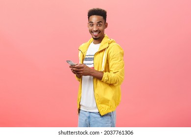 Stylish delighted african american male with afro curly haircut standing half-turned over pink wall holding smartphone wearing yellow trendy jacket smiling joyfully showing friend features of device