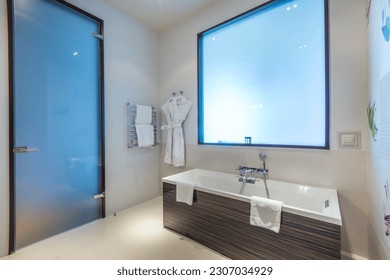 Stylish design of the bathroom in the hotel, with a frosted glass door and a false window. Bath with shower, heated towel rail and bathrobes.