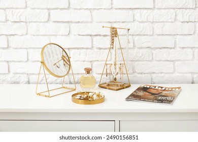 Stylish different jewelry with mirror, perfume bottle and magazine on dresser near white brick wall