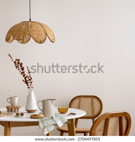 The stylish dining room with round table, rattan chair, lamp and kitchen accessories. Green leaf in vase.  Beige wall. Home decor. Template. 
