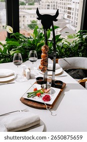 A stylish dining setup features a creative food display with skewered meat, accompanied by fresh vegetables, overlooking a vibrant cityscape through large windows.
