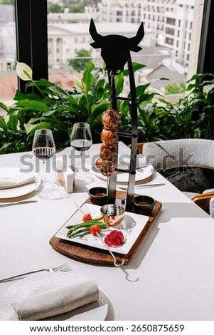 A stylish dining setup features a creative food display with skewered meat, accompanied by fresh vegetables, overlooking a vibrant cityscape through large windows.