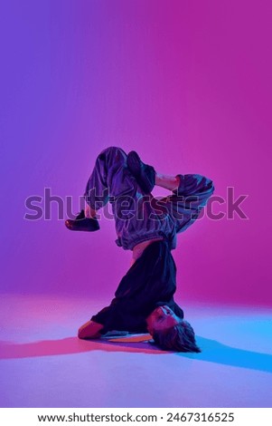Stylish dressed junior dancer, guy performing headstand in motion in mixed neon light against vibrant gradient background. Concept of sport and hobby, music, fashion and art, movement. Ad