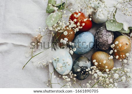 Stylish easter eggs in rustic nest on table flat lay. Natural dyed colorful easter eggs with spring white flowers and feathers on rural textile background. Happy Easter