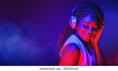 Stylish fashion african american teenager model wearing headphones listening dj music dancing in purple neon lights. Young teen girl enjoy cool music