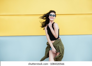Stylish Fashion portrait of trendy cheerful casual young woman in black sunglasses posing near yellow urban wall on windy day with streaming hair. Beauty Lifestyle Happiness Emotions Life People