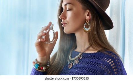 Stylish fashionable attractive brunette boho chic woman with closed eyes wearing jewelry and hat holds perfume bottle 