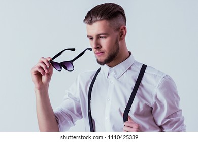 Stylish and fashionable man in classical wear wearing black sunglasses while standing against gray background. Elegant man in white shirt and suspenders posing in studio. Men's classic eyewear fashion