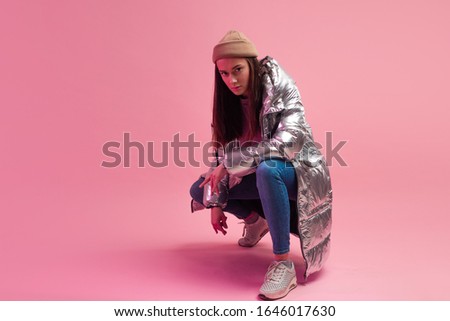 Stylish fashionable and modern young woman in a puffy light down jacket. The jacket is a silver metallic color. Girl in street clothes, jeans and sneakers. Sits on a pink background