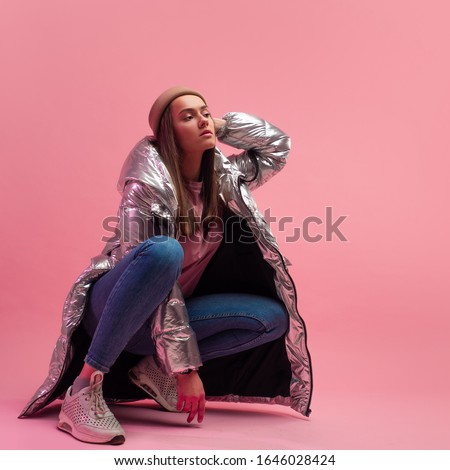 Stylish fashionable and modern young woman in a puffy light down jacket. The jacket is a silver metallic color. Girl in street clothes, jeans and sneakers. Sits on a pink background