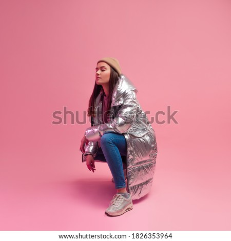 Stylish fashionable and modern young woman in a puffy light down jacket. The jacket is a silver metallic color. woman in street clothes, jeans and sneakers. Sits on a pink background