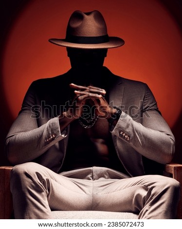 Stylish, fashionable and trendy man in silhouette sitting in a dark room with a red studio background. Edgy, mysterious and curious male mafia boss sitting in a fancy suit, hat and expensive jewelry