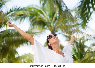 Stylish Fashionable Woman Having Fun And Enjoying Relaxing Summer Tropical Vacation At Beach. Female Brunette Wearing Fashion White Kaftan And Sunglasses Raising Arms.