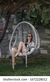 Stylish fashionable woman sitting in hanging rattan egg chair, relaxing summer afternoon