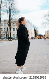 Stylish fashionable young woman in coat holding coffee while walking in park. Back view of caucasian cute woman carelessly looking away outdoors. Vertical view.