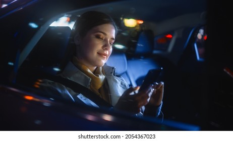 Stylish Female Is Commuting Home In A Backseat Of A Taxi At Night. Beautiful Woman Passenger Using Smartphone While In A Car In Urban City Street With Working Neon Signs.