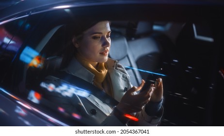 Stylish Female Is Commuting Home In A Backseat Of A Taxi At Night. Beautiful Woman Passenger Using Smartphone While In A Car In Urban City Street With Working Neon Signs.