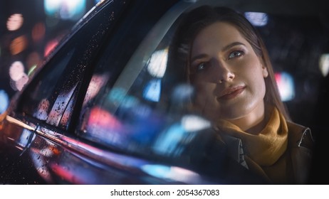 Stylish Female is Commuting Home in a Backseat of a Taxi at Night. Beautiful Woman Passenger Looking Out of Window while in a Car in Urban City Street with Working Neon Signs. - Powered by Shutterstock