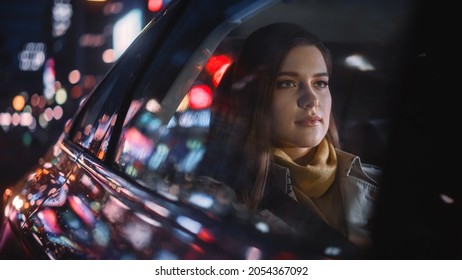 Stylish Female Is Commuting Home In A Backseat Of A Taxi At Night. Beautiful Woman Passenger Looking Out Of Window While In A Car In Urban City Street With Working Neon Signs.