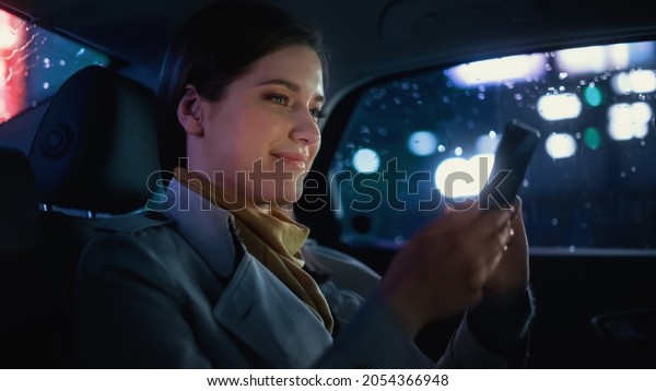 Stylish Female is Commuting Home in a Backseat of\
a Taxi at Night. Beautiful Woman Passenger Using Smartphone and\
Looking Out of Window while in a Car in Urban City Street with\
Working Neon Signs.