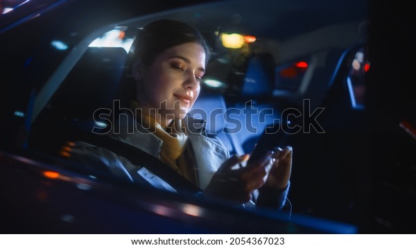 Stylish Female is\
Commuting Home in a Backseat of a Taxi at Night. Beautiful Woman\
Passenger Using Smartphone while in a Car in Urban City Street with\
Working Neon Signs.