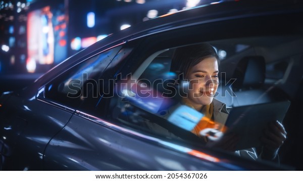 Stylish Female\
is Commuting Home in a Backseat of a Taxi at Night. Beautiful Woman\
Passenger Using Tablet Computer while in Car in Urban City Street\
with Working Neon\
Signs.