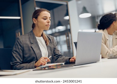 Stylish female manager working on laptop while sitting the desk in office on colleague background - Powered by Shutterstock