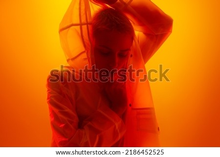 Stylish female model with short blond hair adjusting hood of futuristic raincoat and looking down thoughtfully in bright orange studio with neon light