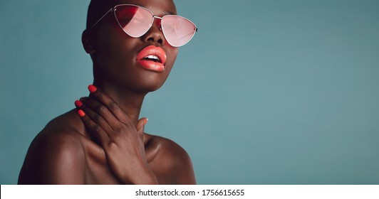 Stylish female model with vibrant makeup wearing trendy sunglasses against grey background. Glamorous woman with red lipstick posing in studio.
